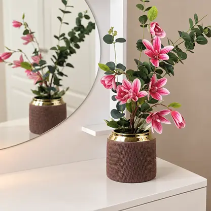 A decorative artificial floral arrangement in a ribbed brown pot with a gold rim, placed on a white vanity table in front of a beige wall and a round mirror.