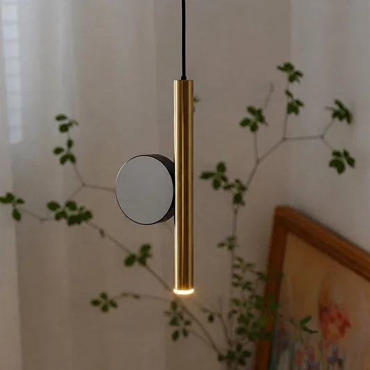 A modern cylindrical pendant light with a brushed gold finish and a circular black accent, shown hanging in a softly lit room with blurred greenery and a framed picture in the background.