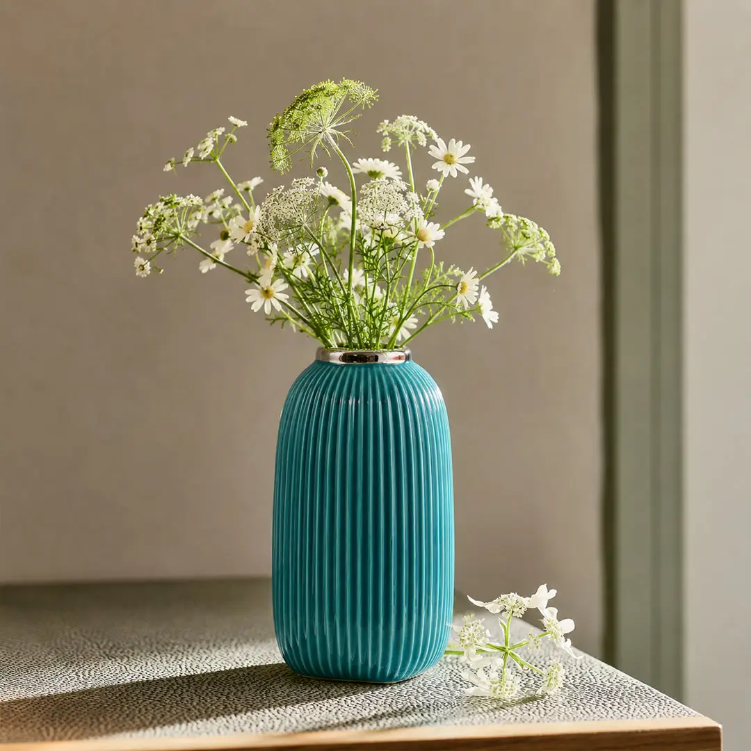 Lifestyle image of a teal fluted ceramic vase styled with white wildflowers on a rustic tabletop with warm sunny shadows.