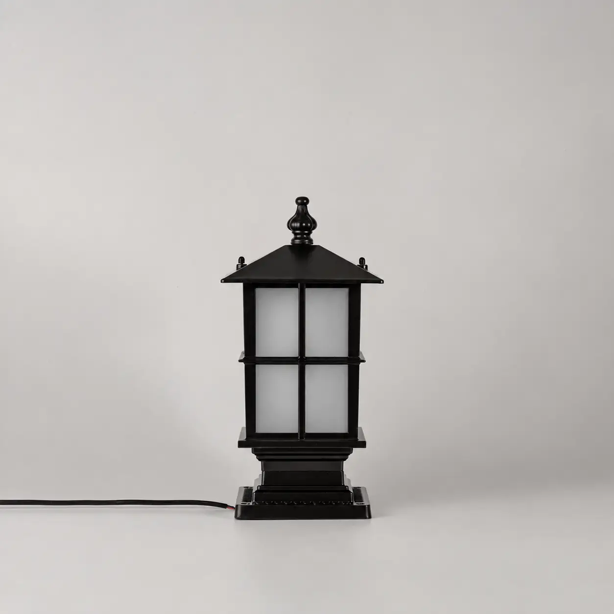 A black outdoor-style lantern light with frosted white glass panels, shown standing on a light gray background with a black power cord extending to the left.