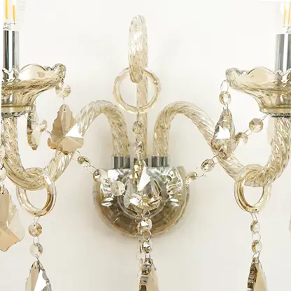 A close-up of a decorative wall sconce with curved clear glass arms, faceted crystal beads, and hanging crystal pendants against a plain light background.