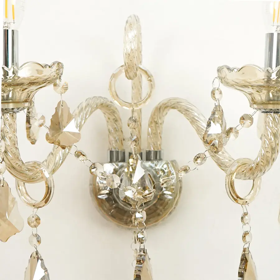 A close-up of a decorative wall sconce with curved clear glass arms, faceted crystal beads, and hanging crystal pendants against a plain light background.
