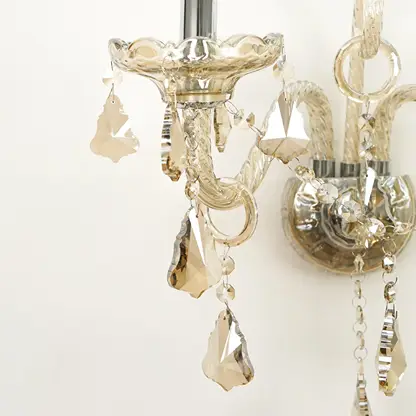 Close-up of a crystal chandelier light fixture showing faceted glass drops and ornate silver-toned metal arms against a plain light background.