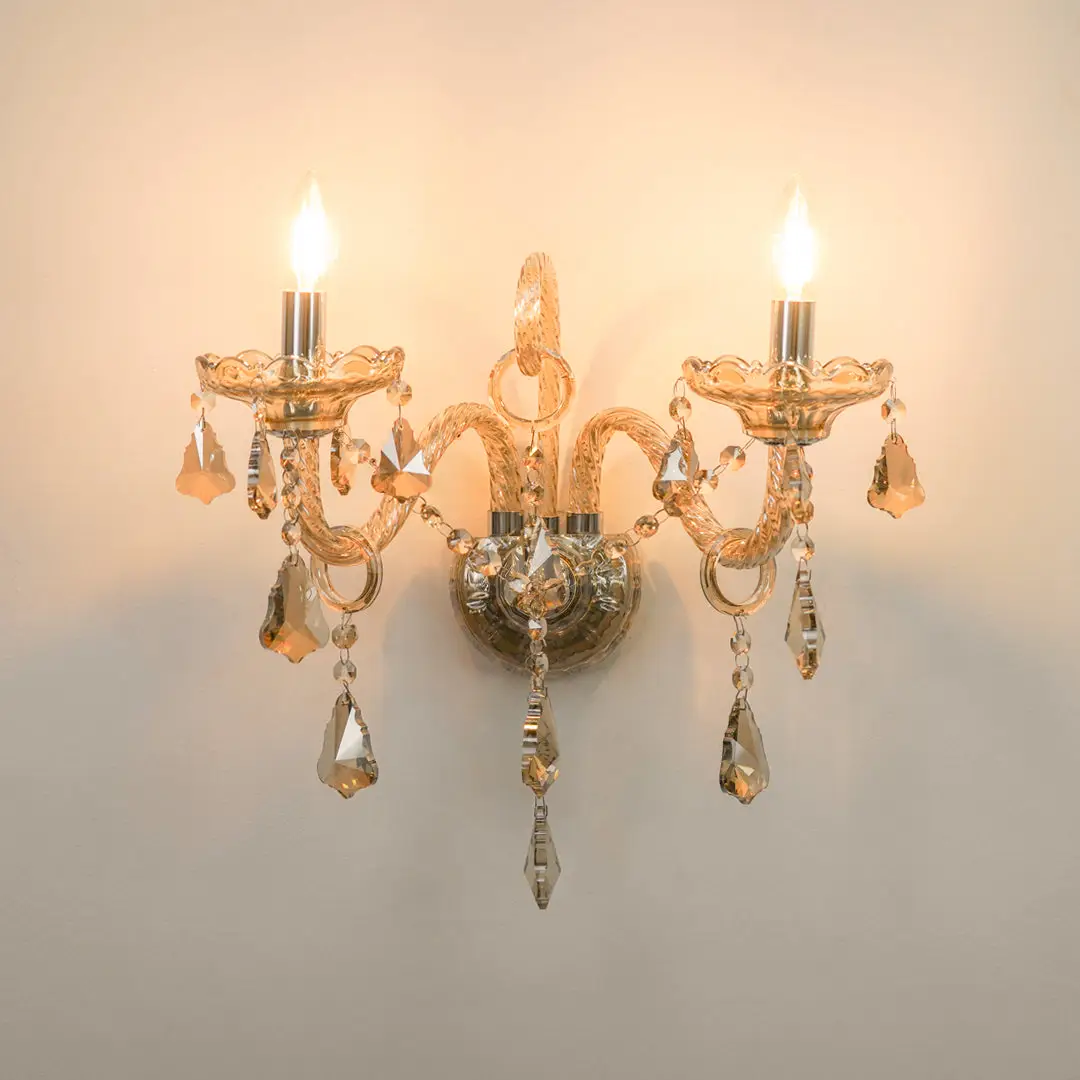 A two-light wall sconce with a warm gold-toned metal frame, ornate curved arms, and hanging clear crystal teardrop pendants, shown lit against a plain light wall.