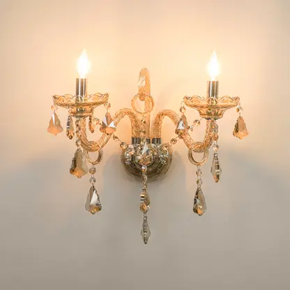 A two-light wall sconce with a warm gold-toned metal frame, ornate curved arms, and hanging clear crystal teardrop pendants, shown lit against a plain light wall.