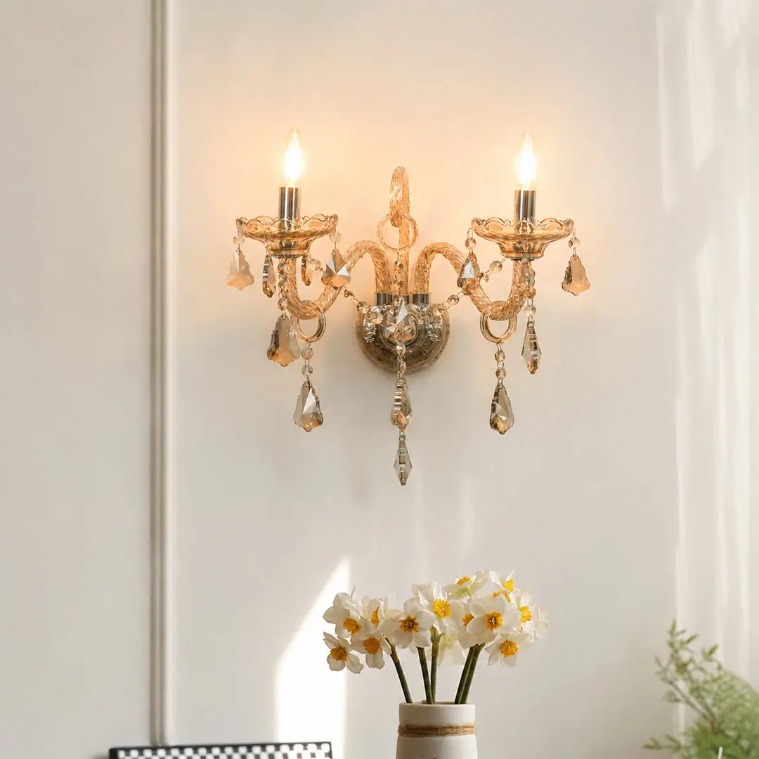 A gold-toned wall sconce with two lit candle-style bulbs and hanging crystal prisms, mounted on a white wall above a vase of white flowers.