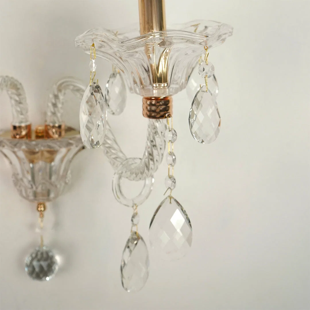A close-up of a clear crystal chandelier detail with faceted teardrop pendants, twisted crystal arms, and a warm brass or gold-toned metal fitting against a plain light background.