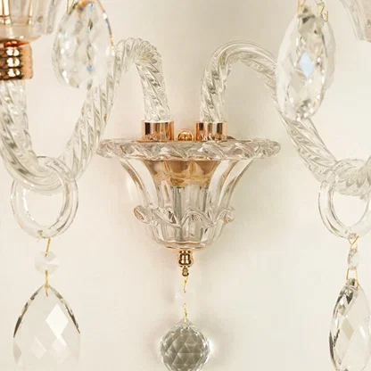 Close-up of a crystal wall sconce with twisted glass arms, clear faceted hanging drops, and warm metallic accents against a light neutral background.