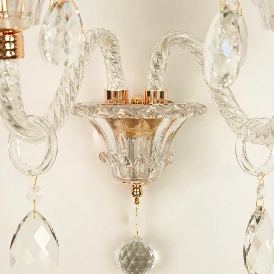 Close-up of a crystal wall sconce with twisted glass arms, clear faceted hanging drops, and warm metallic accents against a light neutral background.