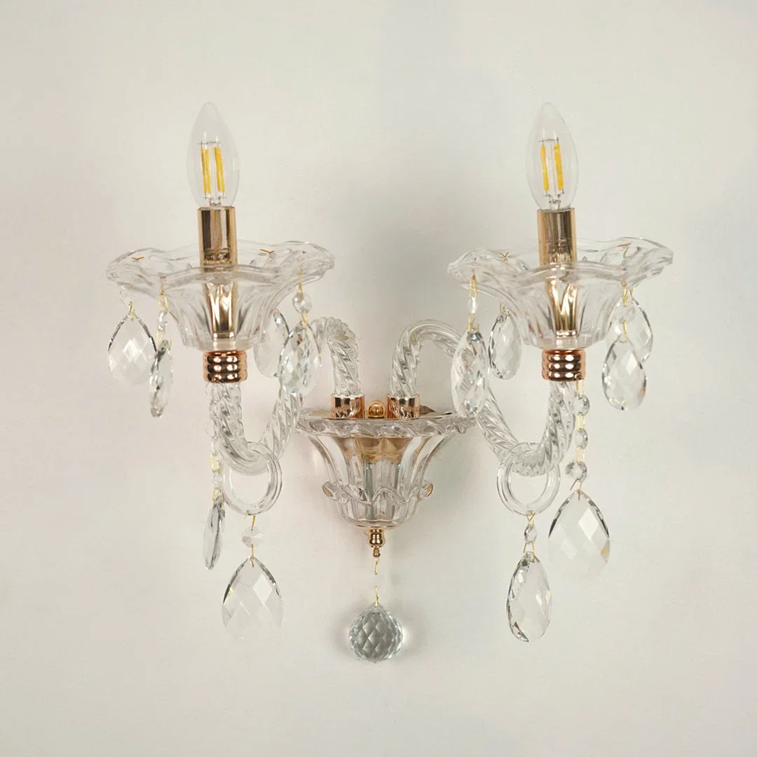 A double-light wall sconce with clear glass crystal prisms and gold-finished metal accents, shown against a plain off-white wall.