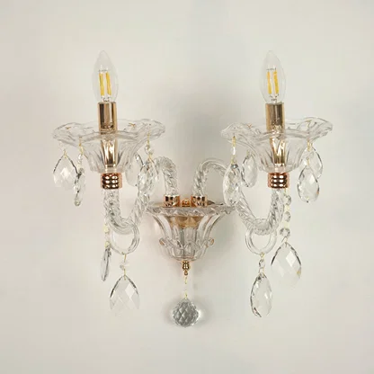 A double-light wall sconce with clear glass crystal prisms and gold-finished metal accents, shown against a plain off-white wall.