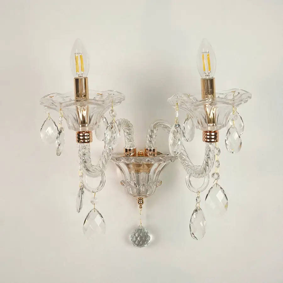 A double-light wall sconce with clear glass crystal prisms and gold-finished metal accents, shown against a plain off-white wall.