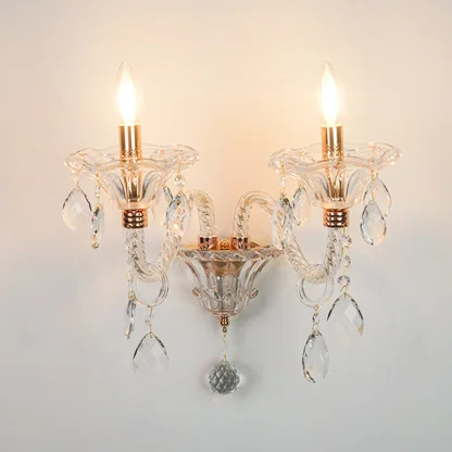 A two-light wall sconce with clear glass crystal arms and hanging teardrop prisms, featuring warm brass/gold-toned accents and illuminated bulbs, mounted against a plain light wall.