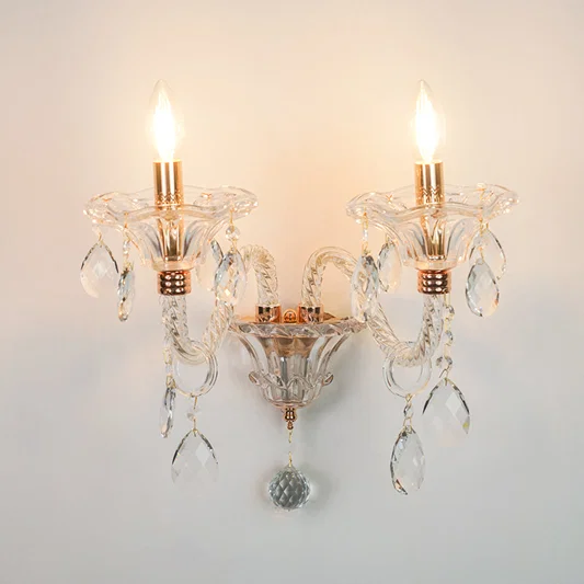A two-light wall sconce with clear glass crystal arms and hanging teardrop prisms, featuring warm brass/gold-toned accents and illuminated bulbs, mounted against a plain light wall.