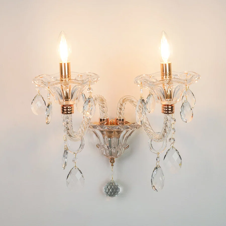 A two-light wall sconce with clear glass crystal arms and hanging teardrop prisms, featuring warm brass/gold-toned accents and illuminated bulbs, mounted against a plain light wall.