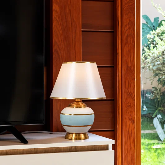 A table lamp with a white conical shade, a light blue and white striped ceramic base, and gold trim, placed on a white console next to a TV in a modern interior setting.