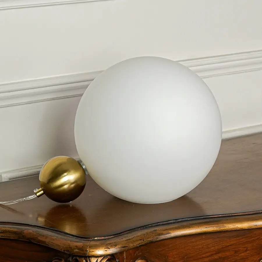 Contemporary matte white globe table lamp with brass finish base positioned on wooden table with neutral wall background ideal for modern lighting setup.
