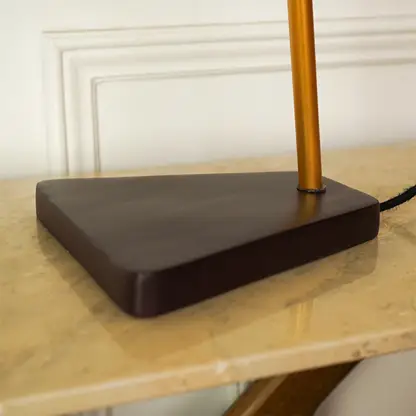 A close-up of a table lamp base with a dark brown triangular metal platform and a vertical gold-toned pole, set on a beige marble tabletop against an off-white paneled wall.