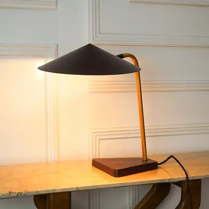 A modern table lamp with a matte black conical shade, a warm orange-brown curved metal stem, and a dark wood base, illuminated on a light marble tabletop against a cream paneled wall.