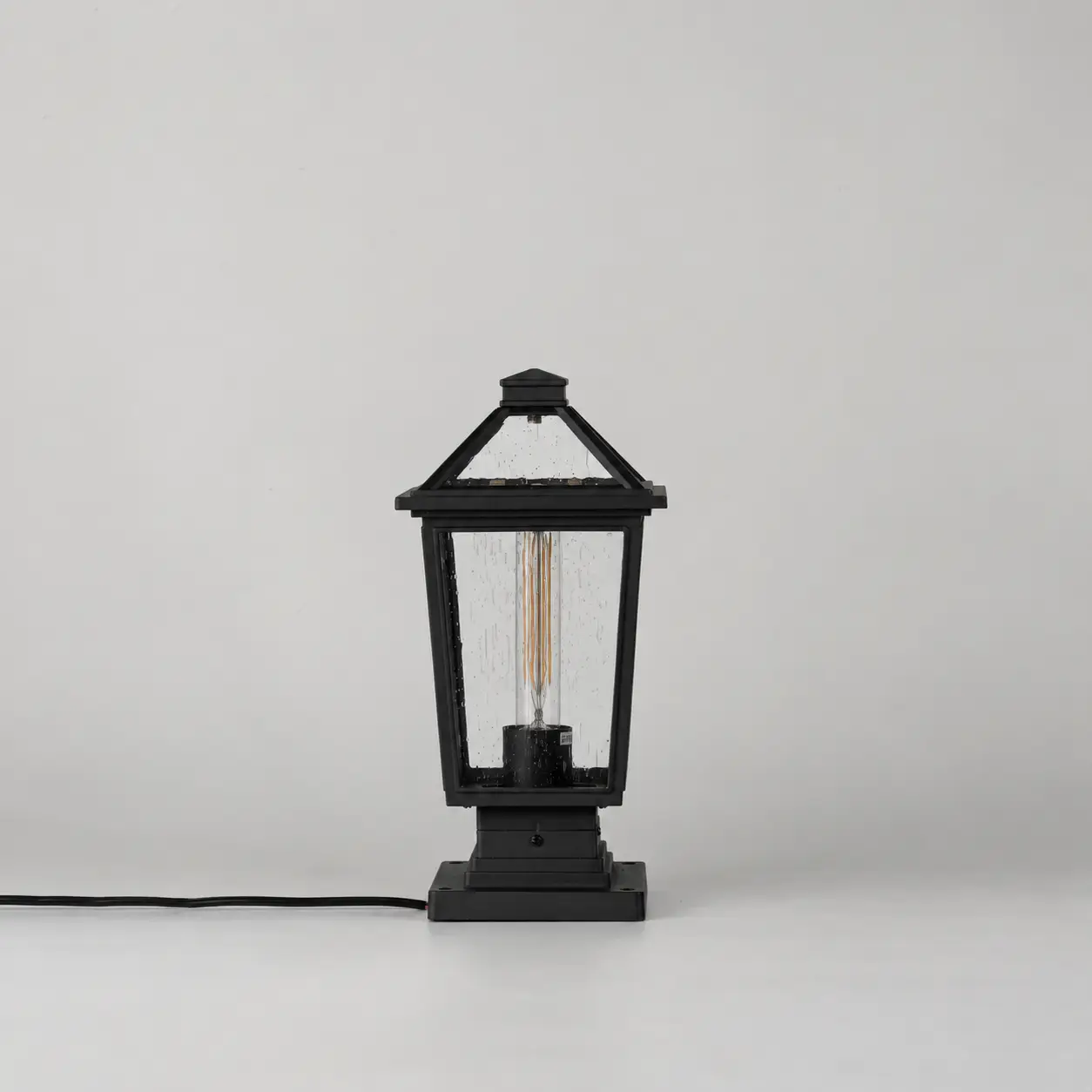 A black lantern-style table lamp with a visible filament bulb inside, shown on a clean light gray studio background. A black power cord extends from the left side of the lamp base.