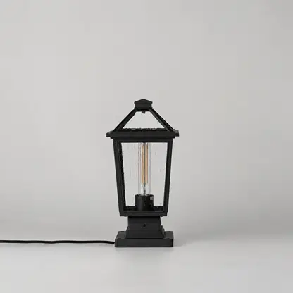 A black lantern-style table lamp with a visible filament bulb inside, shown on a clean light gray studio background. A black power cord extends from the left side of the lamp base.