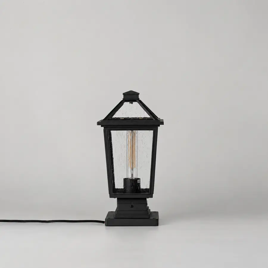 A black lantern-style table lamp with a visible filament bulb inside, shown on a clean light gray studio background. A black power cord extends from the left side of the lamp base.