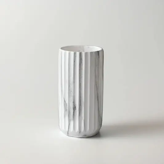 A white ceramic cylindrical cup or tumbler with vertical fluted ridges and a subtle gray marble pattern, set against a plain light gray background.