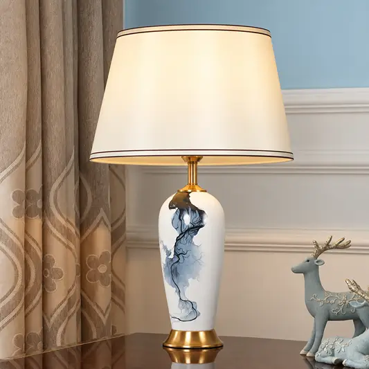 A table lamp with a white ceramic vase-like base featuring a blue and gray abstract painting, a gold-toned neck and base, and a large cream-colored drum shade. It is styled on a dark wooden table next to a blue deer figurine and patterned curtains.