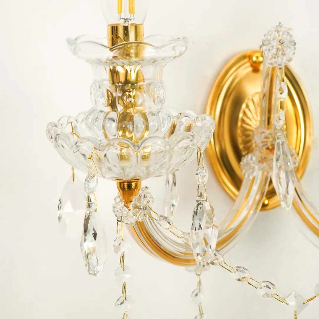 A close-up of a wall-mounted crystal chandelier light fixture with gold-finished metal arms and clear glass prisms hanging against a plain light background.