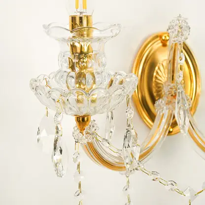 A close-up of a wall-mounted crystal chandelier light fixture with gold-finished metal arms and clear glass prisms hanging against a plain light background.