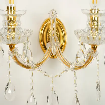 A close-up of a two-light wall sconce with polished gold metal arms, clear glass candle-style holders, and hanging crystal prisms against a plain light background.