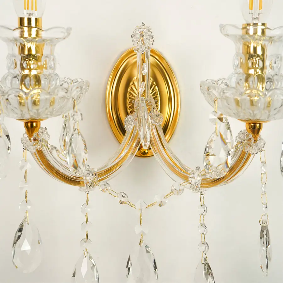 A close-up of a two-light wall sconce with polished gold metal arms, clear glass candle-style holders, and hanging crystal prisms against a plain light background.