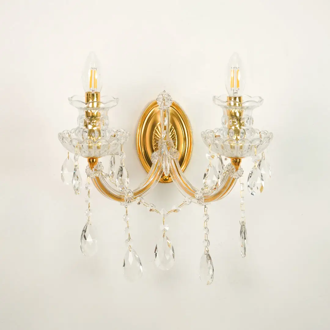 A two-light wall sconce with a polished gold brass frame and clear crystal hanging prisms, shown against a plain off-white wall.