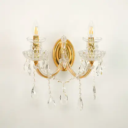 A two-light wall sconce with a polished gold brass frame and clear crystal hanging prisms, shown against a plain off-white wall.