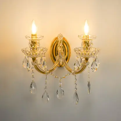 A two-light wall sconce with a polished gold-tone metal frame, clear glass crystal candle-style holders, and hanging teardrop crystal drops, shown illuminated against a plain wall.
