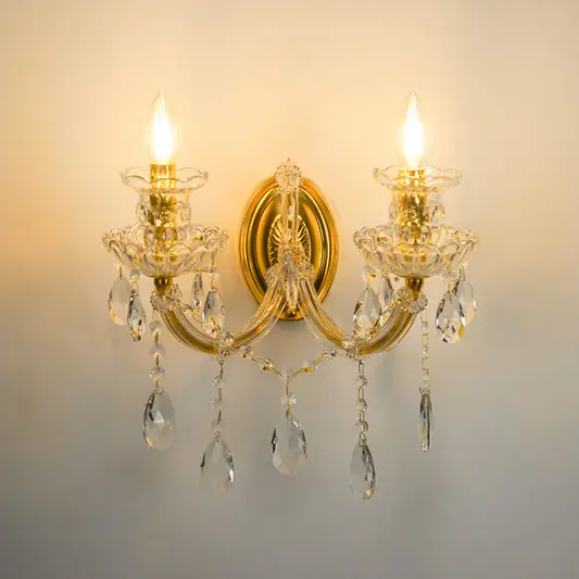 A two-light wall sconce with a polished gold-tone metal frame, clear glass crystal candle-style holders, and hanging teardrop crystal drops, shown illuminated against a plain wall.