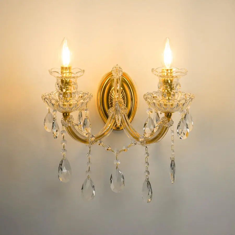 A two-light wall sconce with a polished gold-tone metal frame, clear glass crystal candle-style holders, and hanging teardrop crystal drops, shown illuminated against a plain wall.