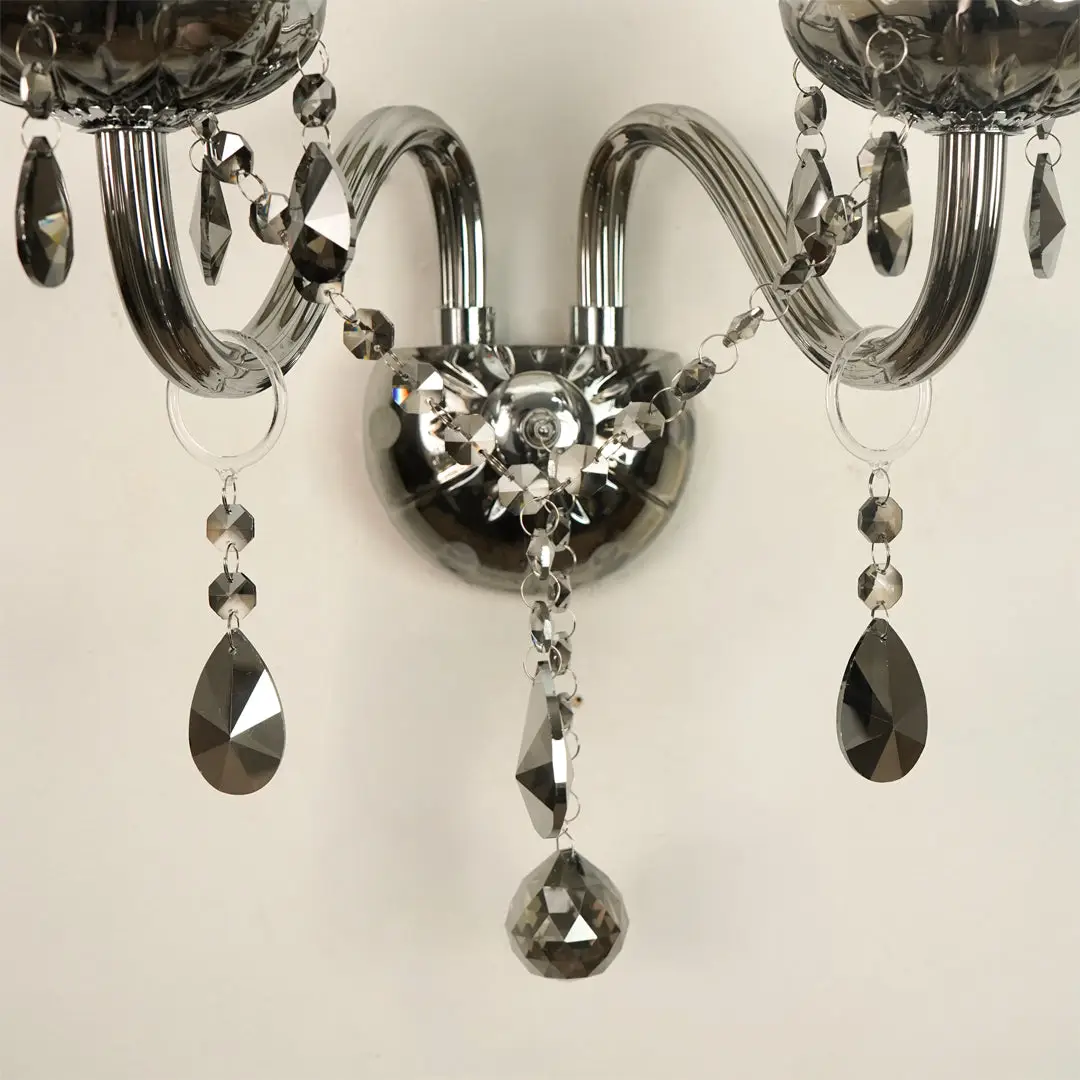 A close-up of a wall-mounted chandelier with curved silver arms, faceted clear crystal beads, and hanging teardrop-shaped dark gray crystal pendants against a plain light background.