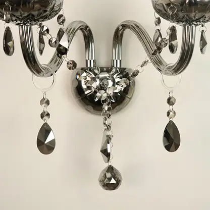 A close-up of a wall-mounted chandelier with curved silver arms, faceted clear crystal beads, and hanging teardrop-shaped dark gray crystal pendants against a plain light background.