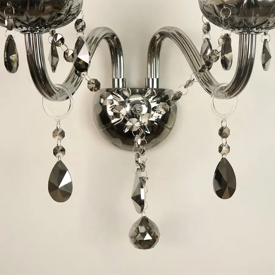 A close-up of a wall-mounted chandelier with curved silver arms, faceted clear crystal beads, and hanging teardrop-shaped dark gray crystal pendants against a plain light background.