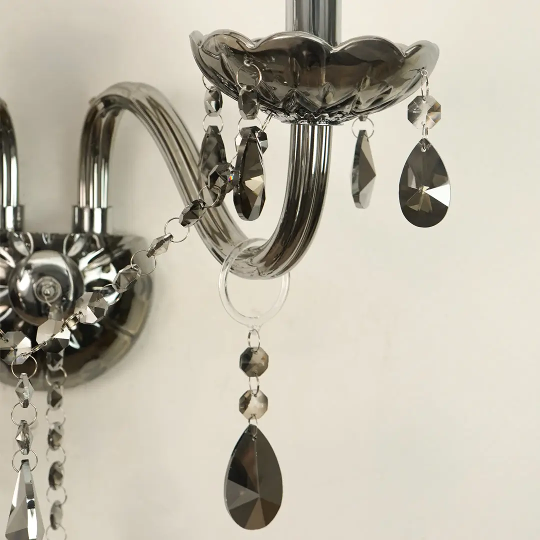 A close-up of a wall-mounted crystal chandelier with polished silver metal arms and faceted teardrop crystal drops in smoky gray and clear glass.