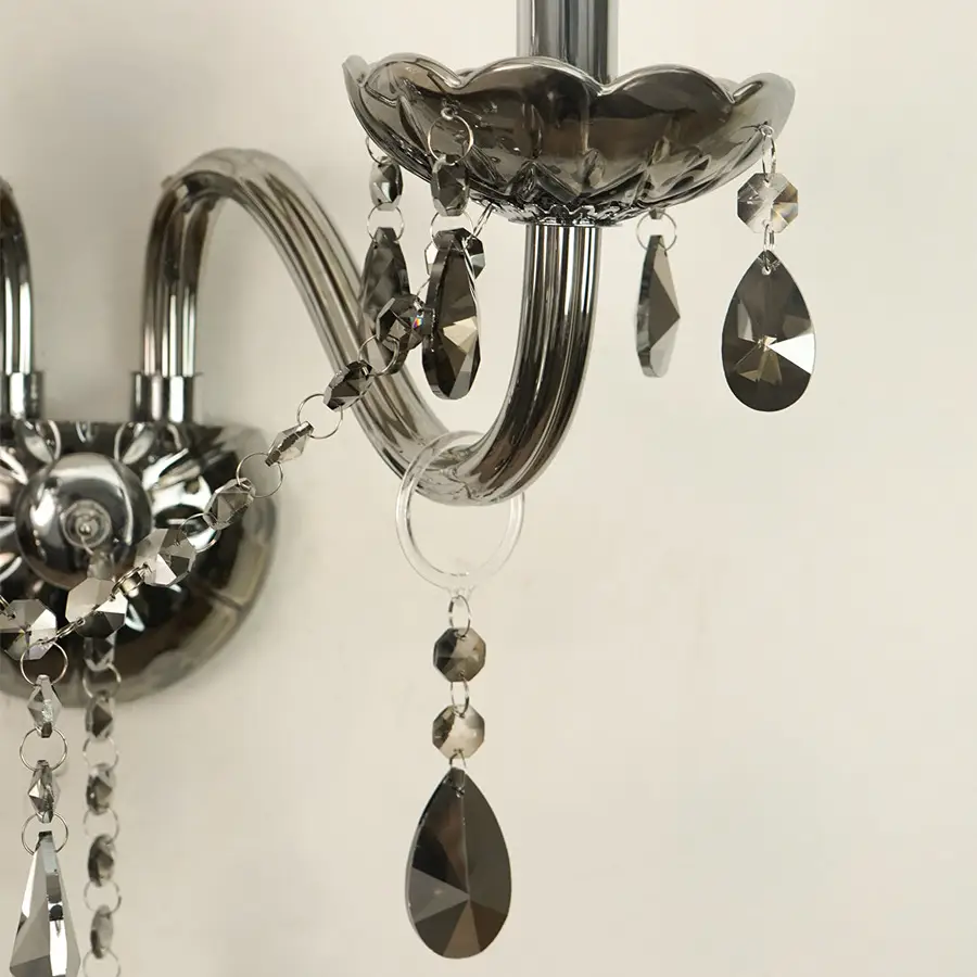 A close-up of a wall-mounted crystal chandelier with polished silver metal arms and faceted teardrop crystal drops in smoky gray and clear glass.