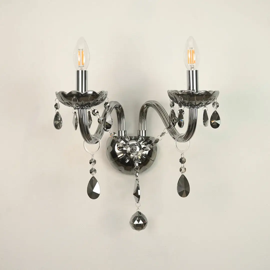 A two-light wall sconce with curved arms, a polished silver-toned metal body, and hanging faceted crystal prisms, shown against a plain off-white wall.
