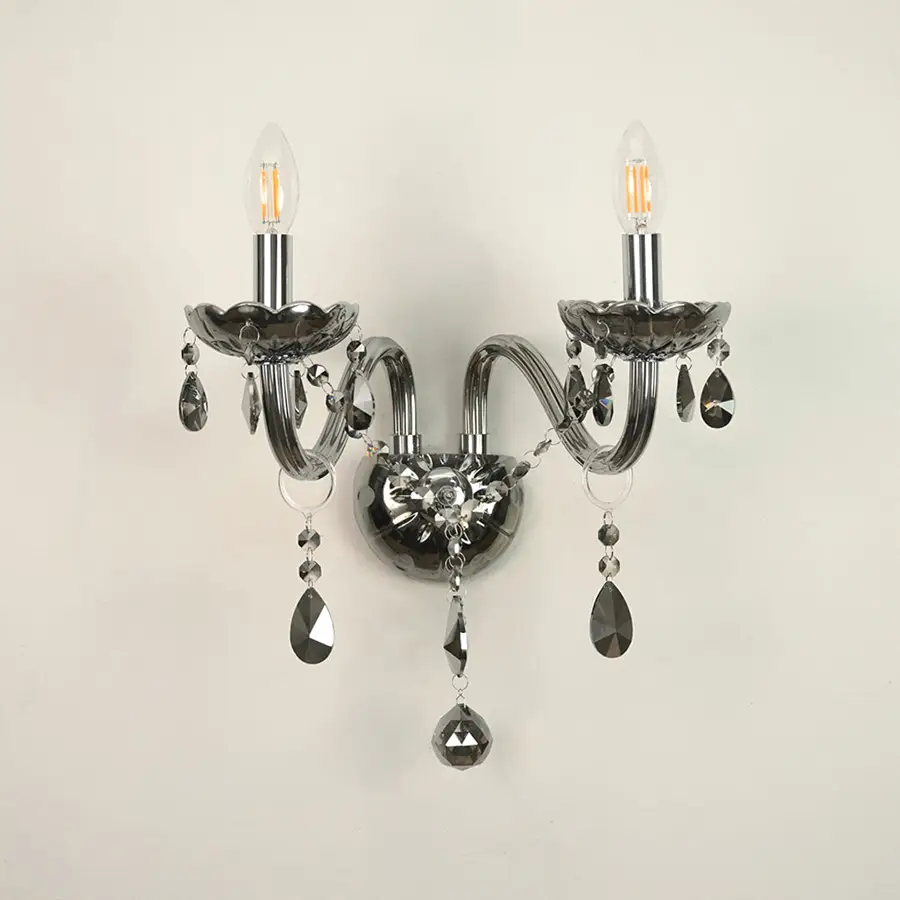 A two-light wall sconce with curved arms, a polished silver-toned metal body, and hanging faceted crystal prisms, shown against a plain off-white wall.