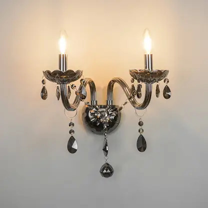 A two-light wall sconce with curved silver-toned metal arms, clear faceted crystal teardrop pendants, and lit candle-style bulbs, mounted against a plain light wall.
