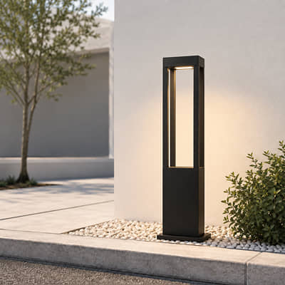 A modern black outdoor bollard light with a rectangular open-top design, illuminated and set on a stone bed next to a concrete walkway and greenery.