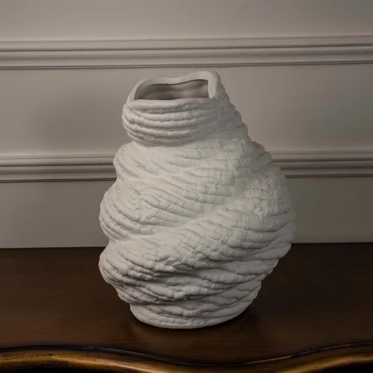 Twisted white ceramic vase with organic textured finish placed on wooden table against neutral wall background showcasing contemporary artistic home decor styling.