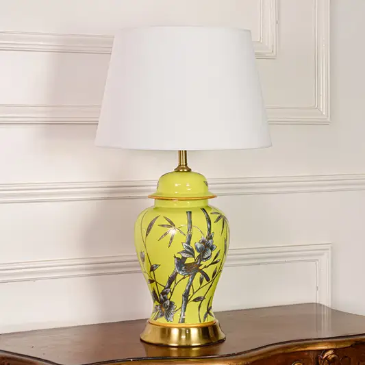 Decorative yellow ceramic table lamp with bamboo bird design gold metal base and fabric shade on off white backdrop.