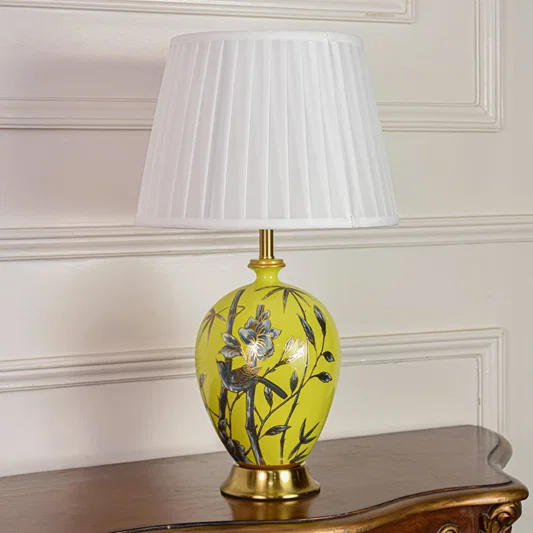 Decorative yellow ceramic table lamp with floral bird artwork gold metal base and pleated fabric shade on off white background.