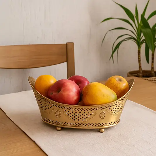 A decorative golden fruit basket filled with fresh apples and mangoes placed on a wooden dining table with a neutral table runner creating a warm and homely lifestyle setting.